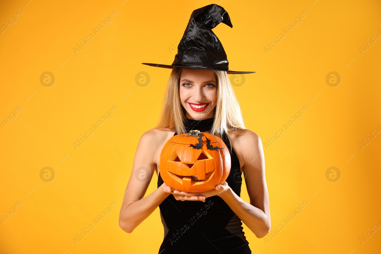 Photo of Beautiful woman in witch costume with jack o'lantern on yellow background. Halloween party