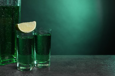 Photo of Absinthe in shot glasses and lime wedge on gray textured table against green background, space for text. Alcoholic drink