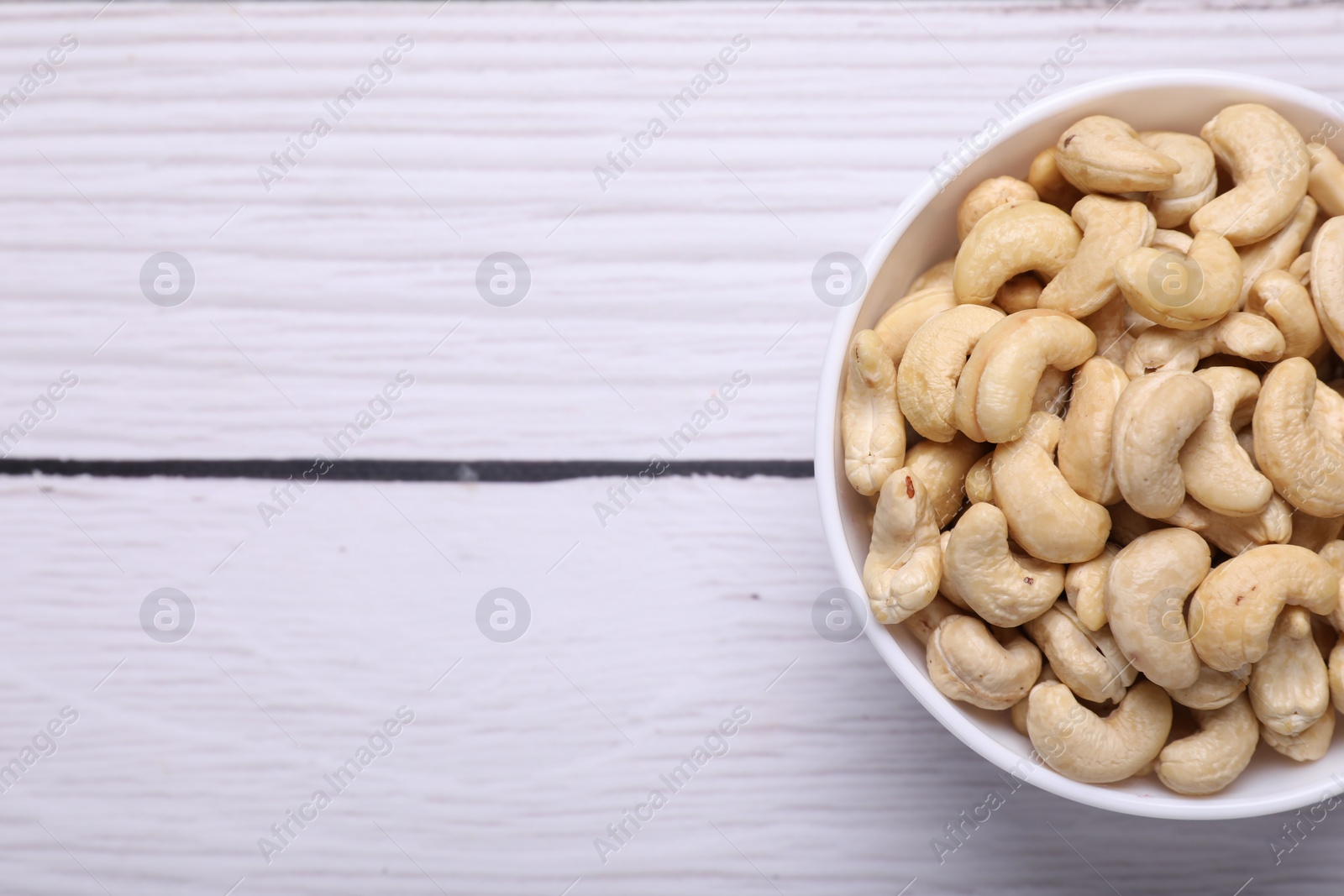 Photo of Tasty cashew nuts in bowl on white wooden table, top view. Space for text