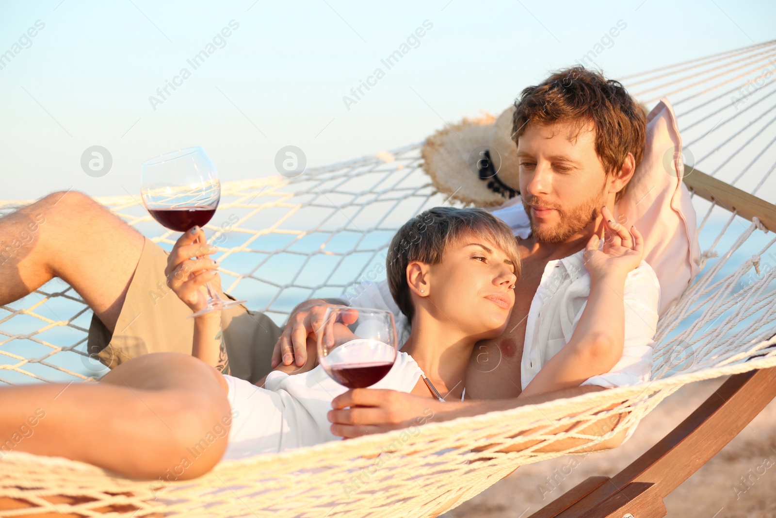 Photo of Young couple resting with glasses of wine in hammock on beach