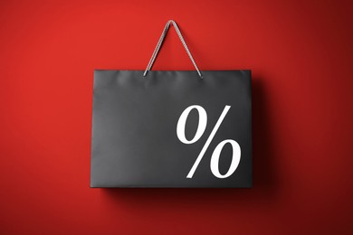 Image of Black paper shopping bag with percent sign hanging on red wall. Discount concept