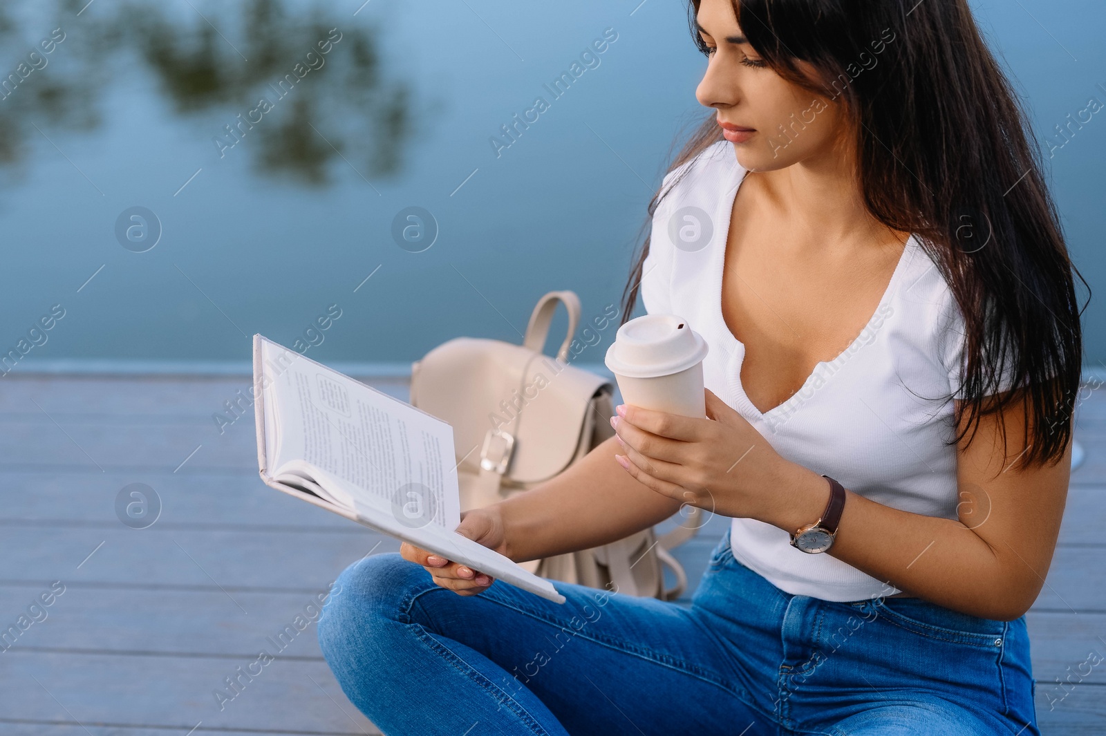 Photo of Young woman with cup of coffee reading book on pier near lake