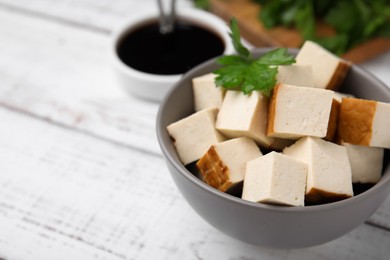 Photo of Bowl of smoked tofu cubes, soy sauce and parsley on white wooden table, closeup. Space for text