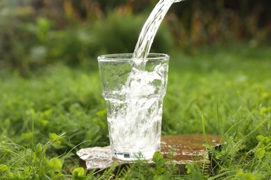 Photo of Pouring fresh water into glass on stone in green grass outdoors