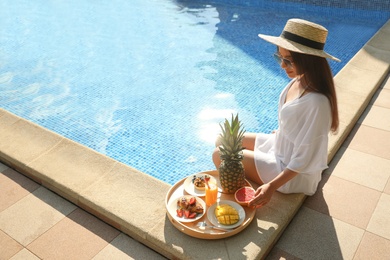 Young woman with delicious breakfast on tray near swimming pool. Space for text