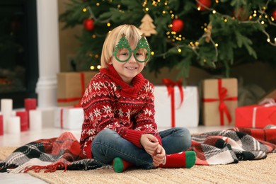 Photo of Little child with plaid and funny glasses on floor near Christmas tree at home
