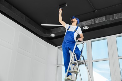 Photo of Worker in uniform painting ceiling with roller indoors, low angle view