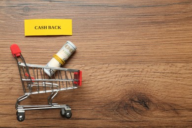 Card with word Cashback, rolled dollar banknotes and shopping cart on wooden background, flat lay. Space for text