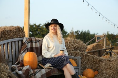 Photo of Beautiful woman with cup of hot drink sitting on wooden bench near hay bales and pumpkins outdoors. Autumn season