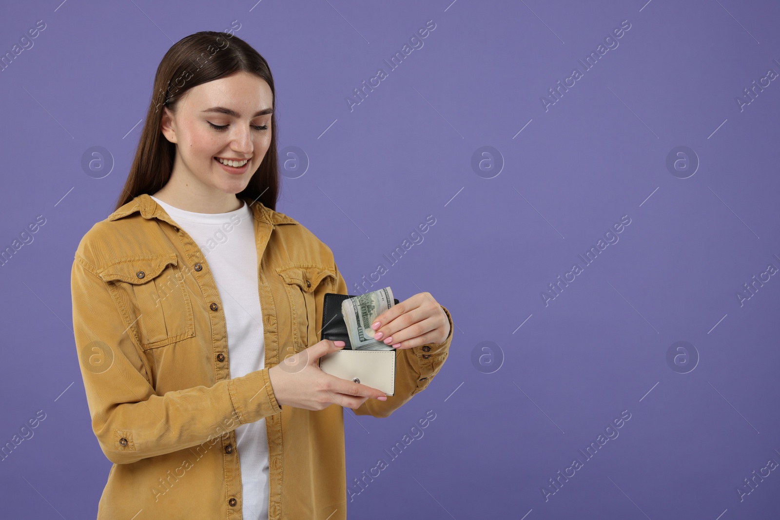 Photo of Happy woman putting dollar banknotes into wallet on purple background, space for text