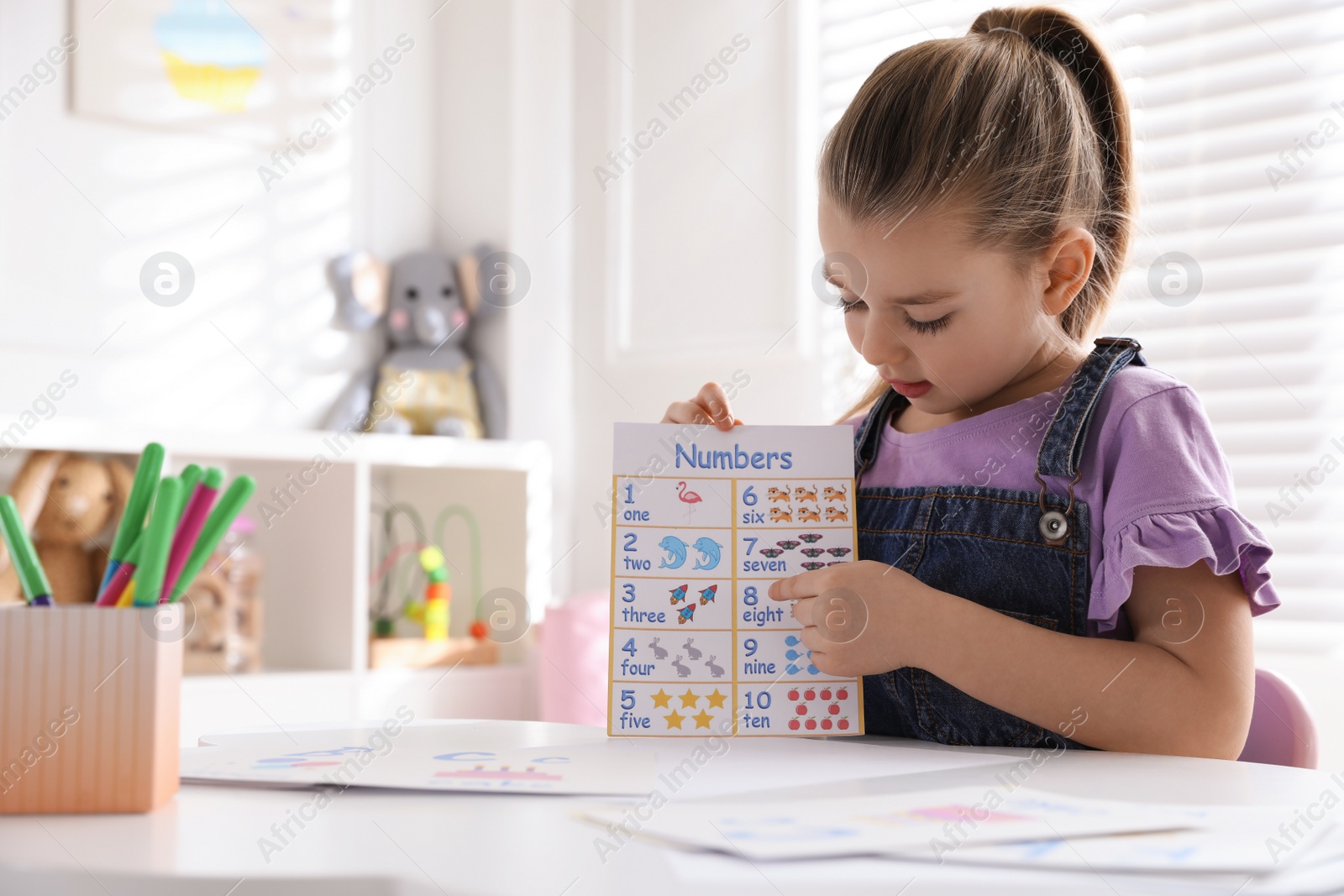 Photo of Little girl holding card with numbers in classroom at English lesson