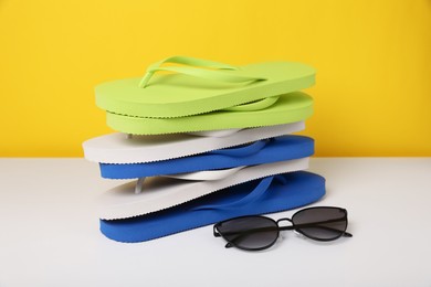 Stack of different flip flops and sunglasses on white table against yellow background