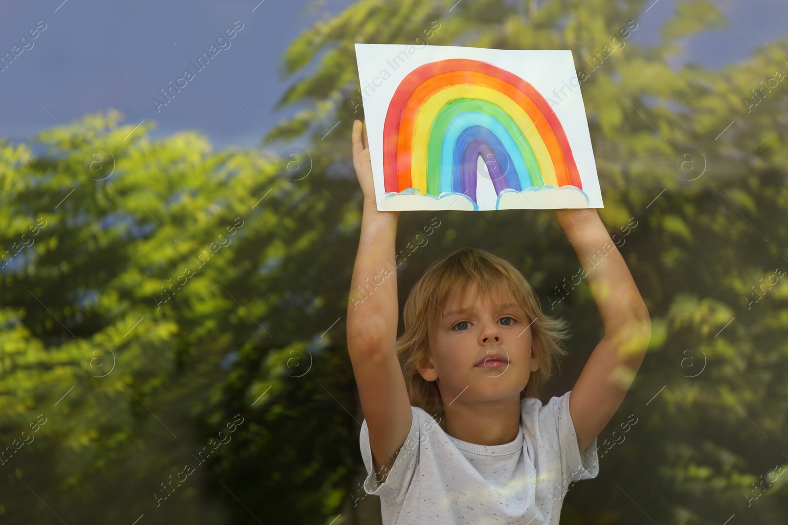 Photo of Little boy with picture of rainbow near window, view from outdoors. Stay at home concept