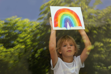 Little boy with picture of rainbow near window, view from outdoors. Stay at home concept