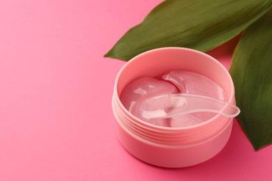 Photo of Jar of under eye patches with spoon and green leaves on pink background, closeup. Space for text