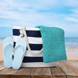 Image of Stylish beach bag, flip flops and towel on wooden surface near seashore 