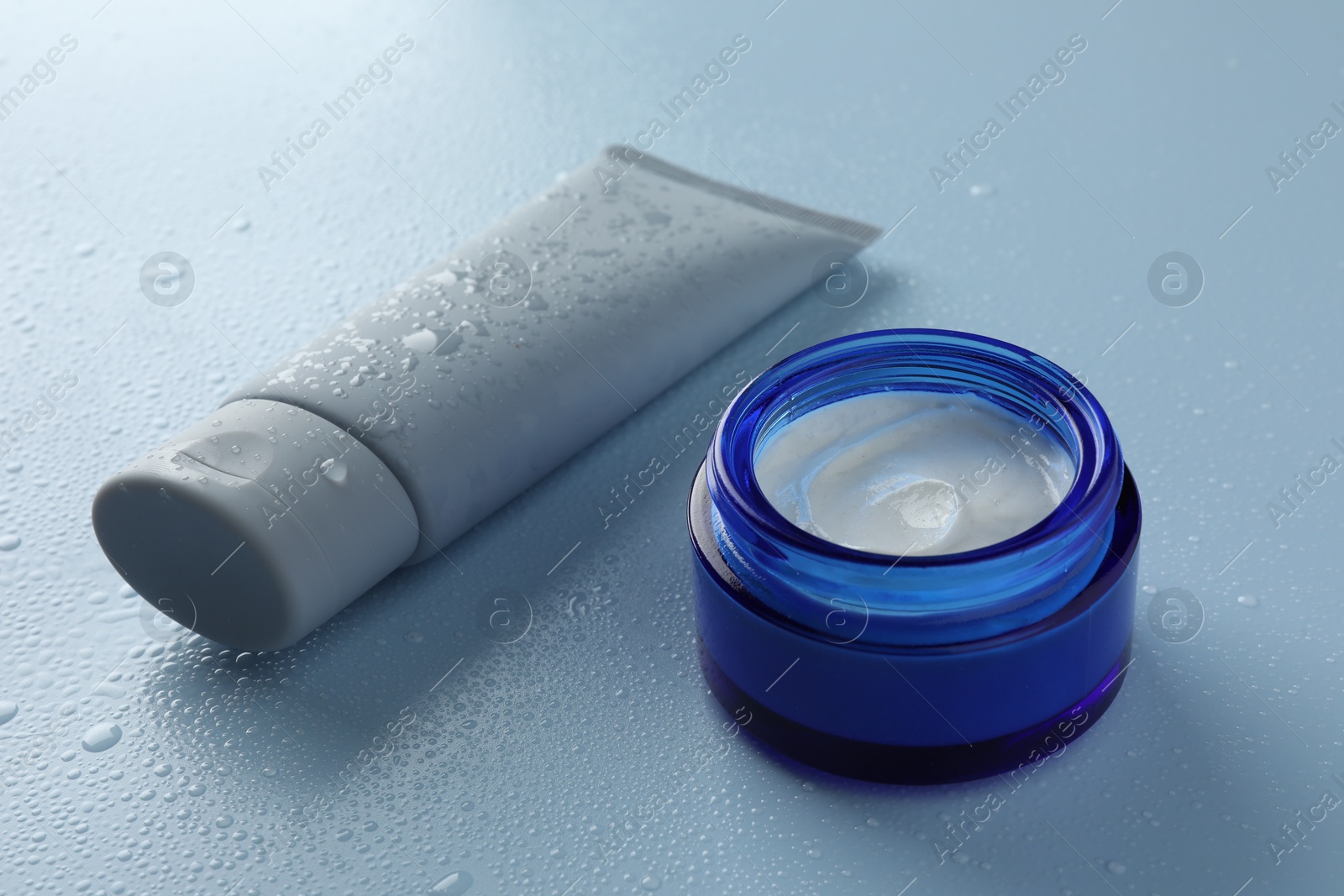 Photo of Moisturizing cream in tube and jar on light blue background with water drops, closeup