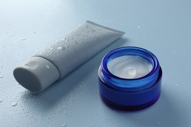Photo of Moisturizing cream in tube and jar on light blue background with water drops, closeup