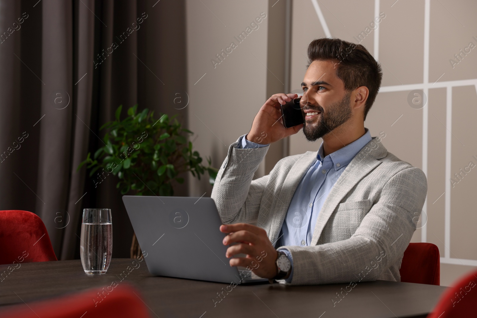 Photo of Happy man using modern laptop while talking on smartphone at table in office