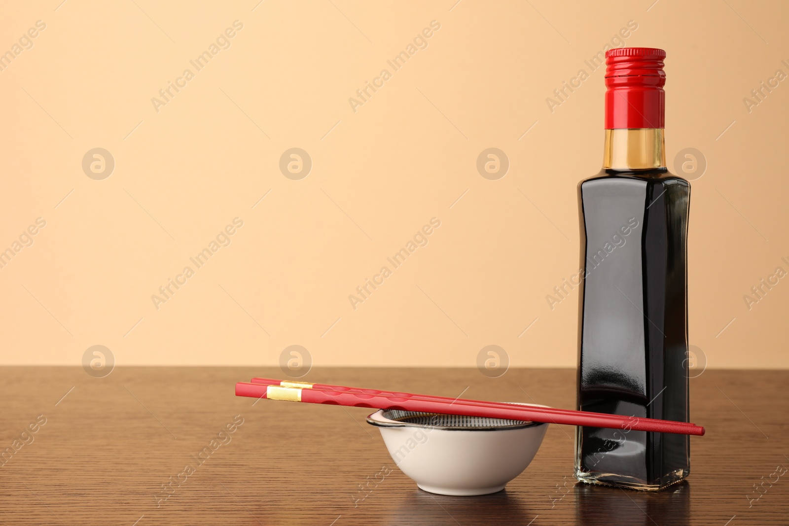 Photo of Bottle, bowl with soy sauce and chopsticks on wooden table against beige background. Space for text