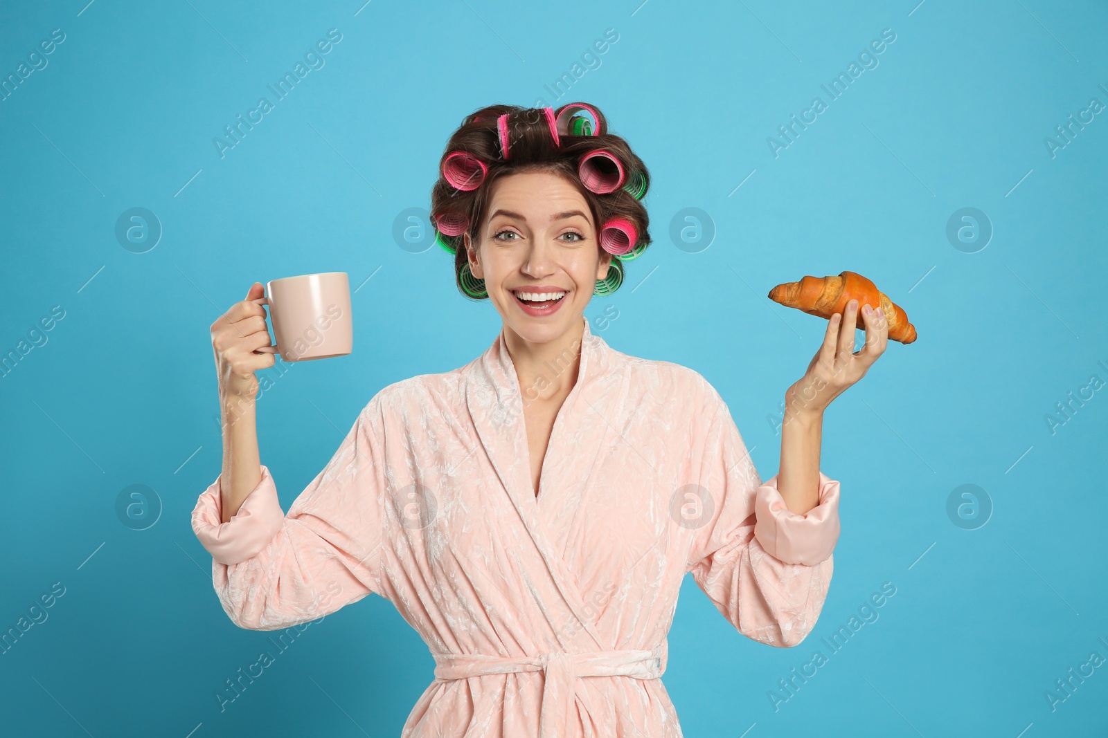 Photo of Happy young woman in bathrobe with hair curlers holding croissant and cup of drink on light blue background