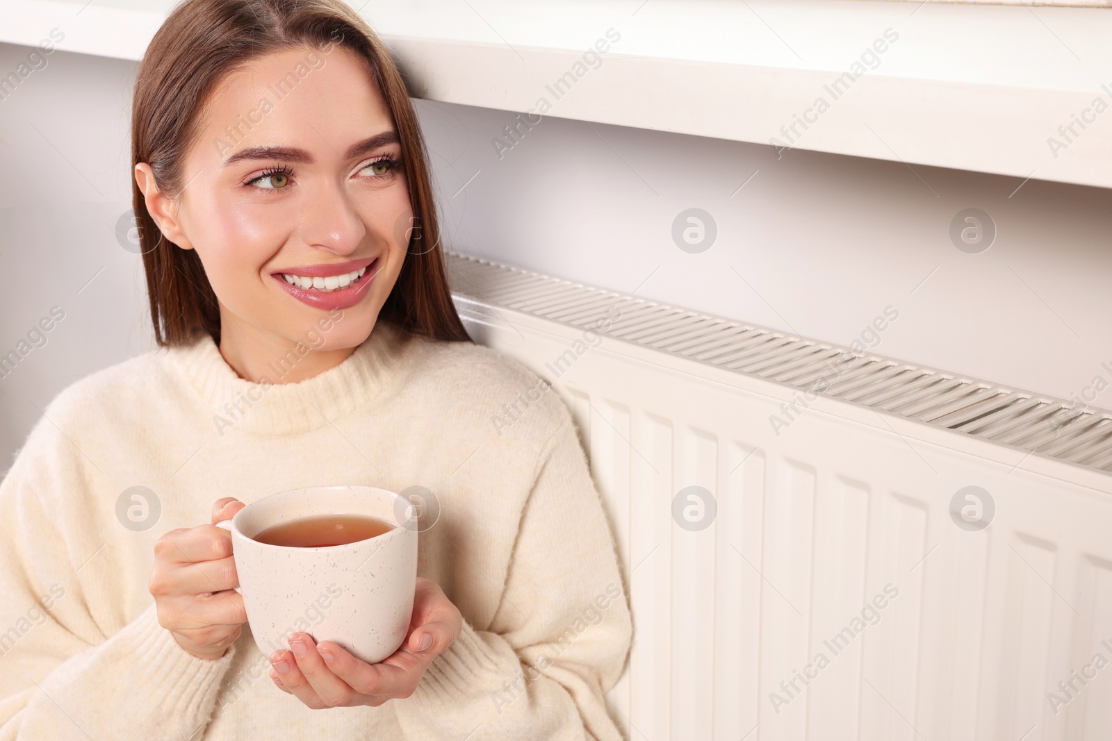 Photo of Woman holding cup with hot drink near heating radiator indoors