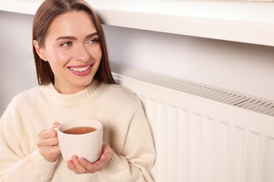 Photo of Woman holding cup with hot drink near heating radiator indoors