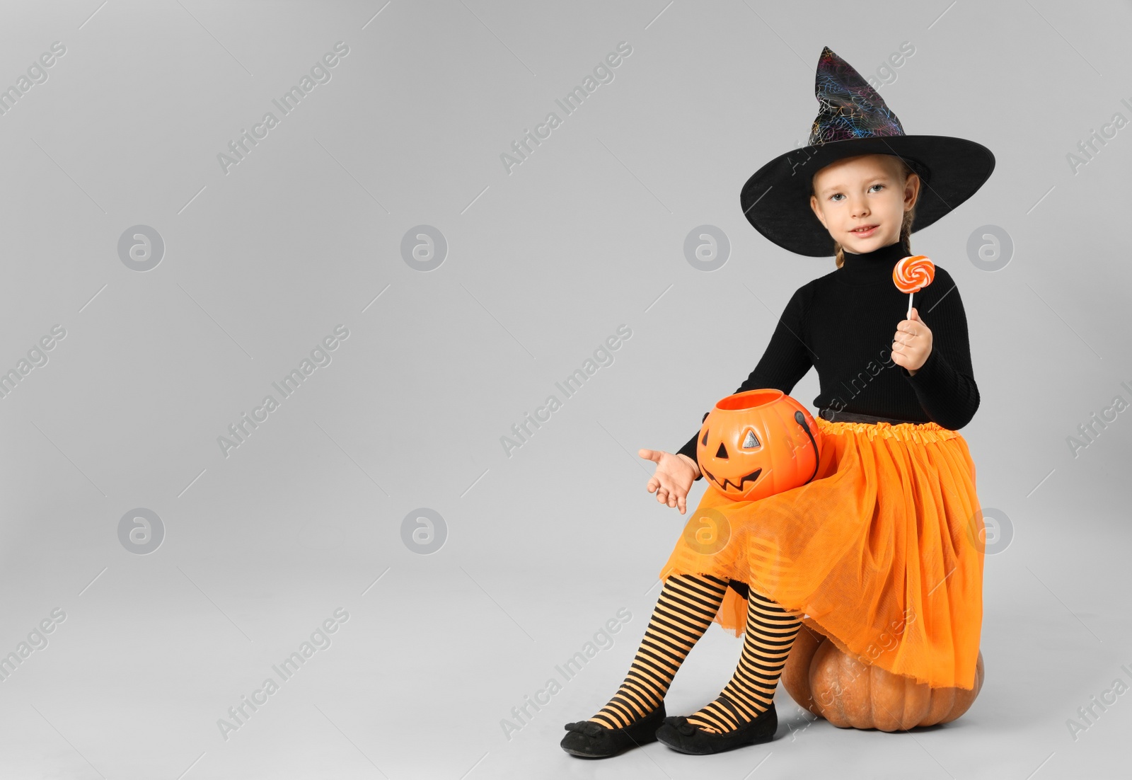 Photo of Cute little girl with pumpkin candy bucket and lollipop wearing Halloween costume on grey background. Space for text