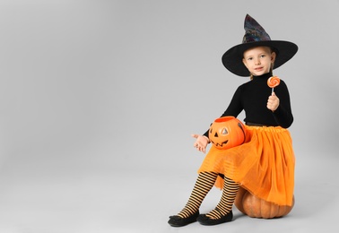 Cute little girl with pumpkin candy bucket and lollipop wearing Halloween costume on grey background. Space for text