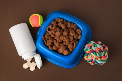 Bowl with dry pet food, bottle of vitamins and toys on brown background, flat lay