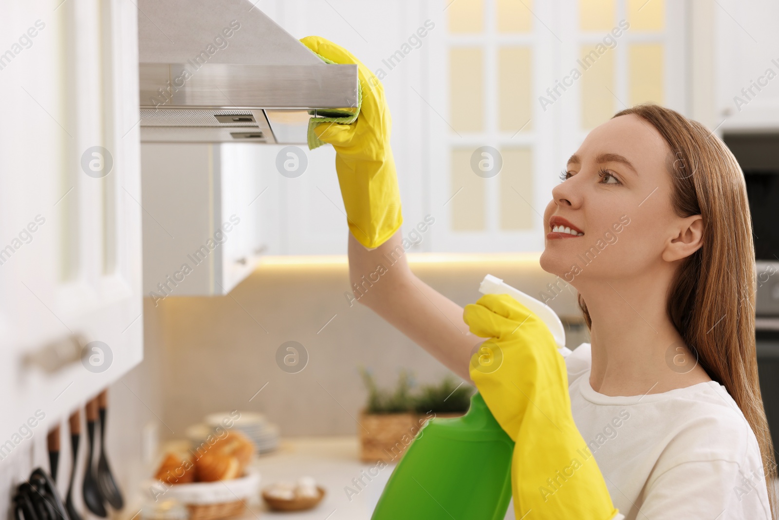 Photo of Woman with spray bottle and microfiber cloth cleaning kitchen hood at home