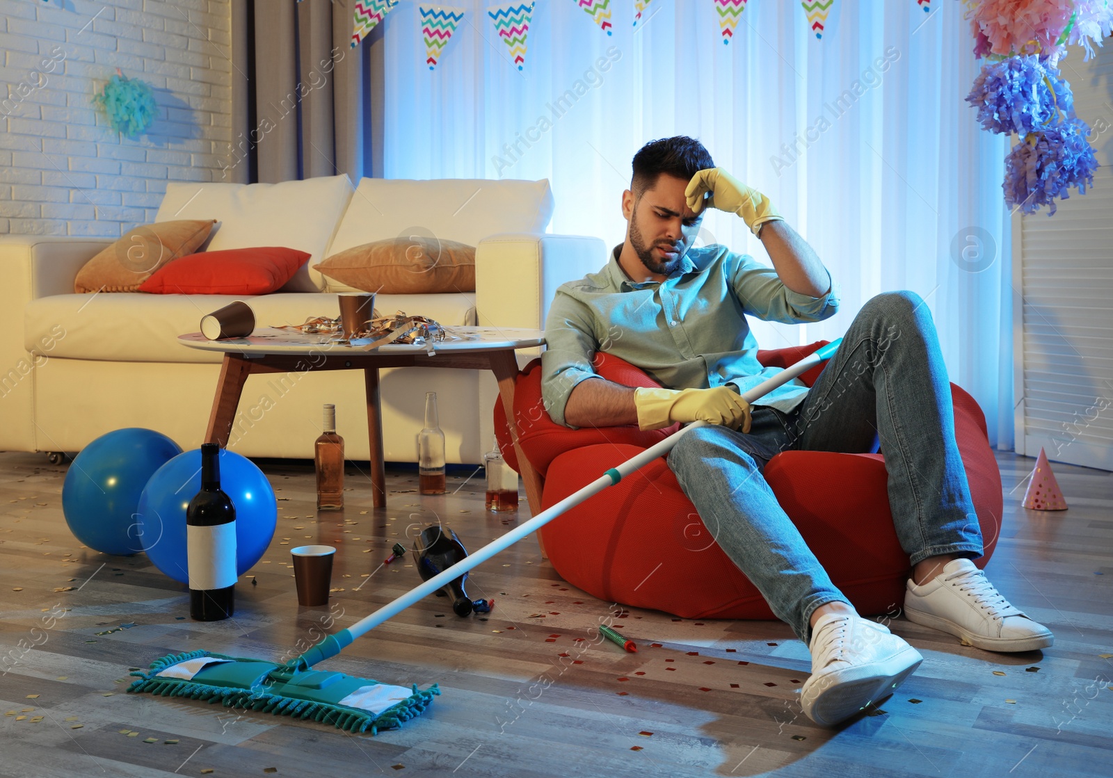 Photo of Young man with mop suffering from hangover in messy room after party