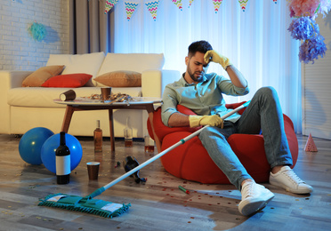 Young man with mop suffering from hangover in messy room after party