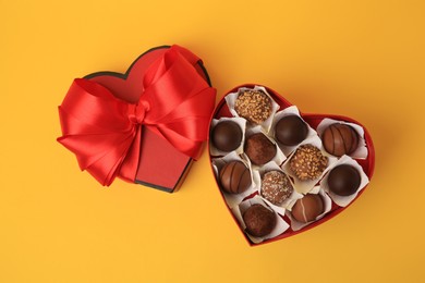 Photo of Heart shaped box with delicious chocolate candies on yellow background, flat lay