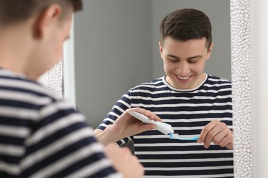 Photo of Happy man squeezing toothpaste from tube onto toothbrush near mirror in bathroom