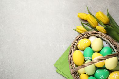 Photo of Many Easter eggs in wicker basket and yellow tulips on grey background, flat lay. Space for text