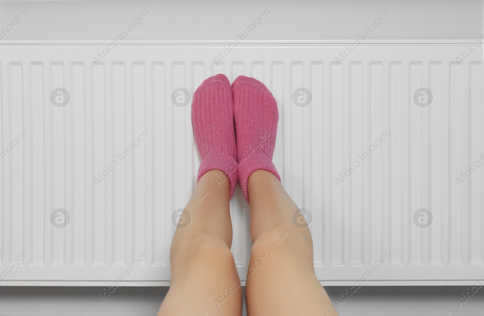 Photo of Woman warming legs on heating radiator near white wall, closeup