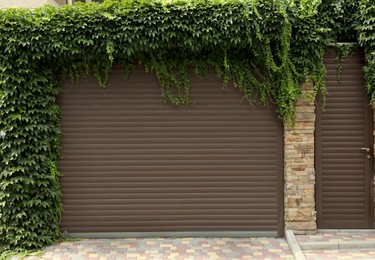 Photo of Brown roller shutter door of garage near entrance overgrown with beautiful climbing plants
