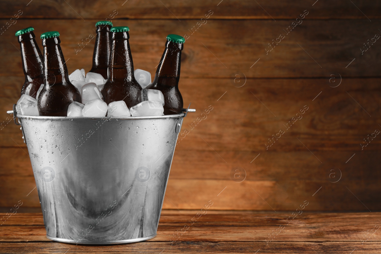 Photo of Metal bucket with bottles of beer and ice cubes on wooden table, space for text