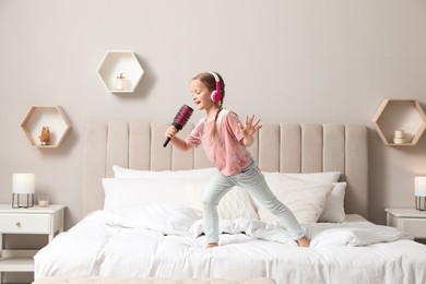Photo of Cute little girl in headphones with hairbrush singing on bed at home