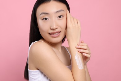 Photo of Beautiful young Asian woman applying body cream onto arm on pink background