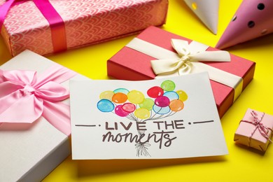 Photo of Card with life-affirming phrase Live The Moments and gift boxes on yellow background