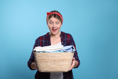 Emotional woman with basket full of laundry on light blue background