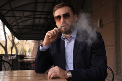 Photo of Handsome young businessman using disposable electronic cigarette at table in outdoor cafe