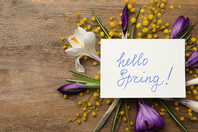 Photo of Card with words HELLO SPRING and fresh flowers on wooden table, flat lay