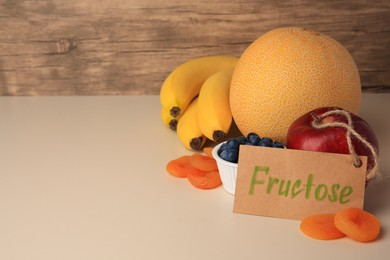 Photo of Card with word Fructose, delicious ripe fruits, blueberries and dried apricots on beige table, space for text