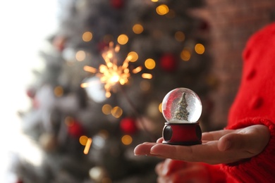 Woman with snow globe and sparkler near Christmas tree, closeup. Space for text