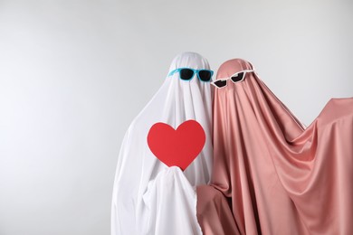 Photo of Cute ghosts. Couple in color sheets and sunglasses with red heart on light grey background, space for text