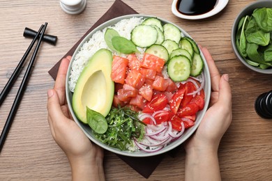 Photo of Woman holding delicious poke bowl with salmon and vegetables at wooden table, top view