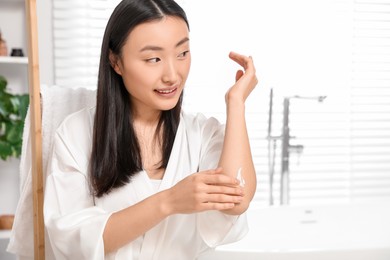 Photo of Beautiful young Asian woman applying body cream on elbow in bathroom, space for text